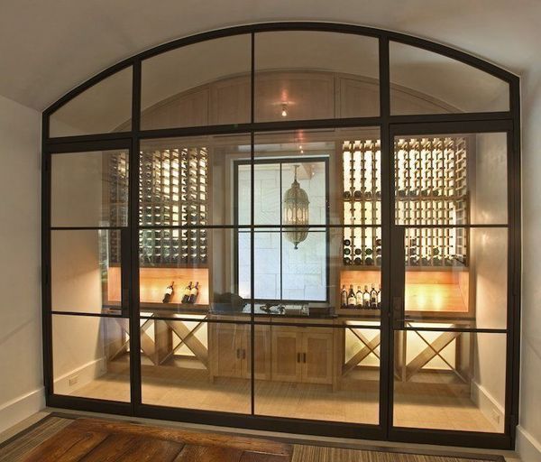 Wine cellar behind a large glass and black metal frame. Wooden wine racks, shelves, and a cabinet are visible.
