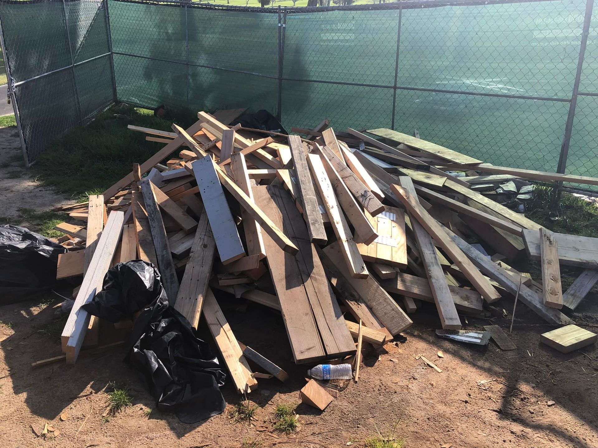 Pile of wooden planks and boards, possibly construction debris, in front of a chain-link fence and green netting.