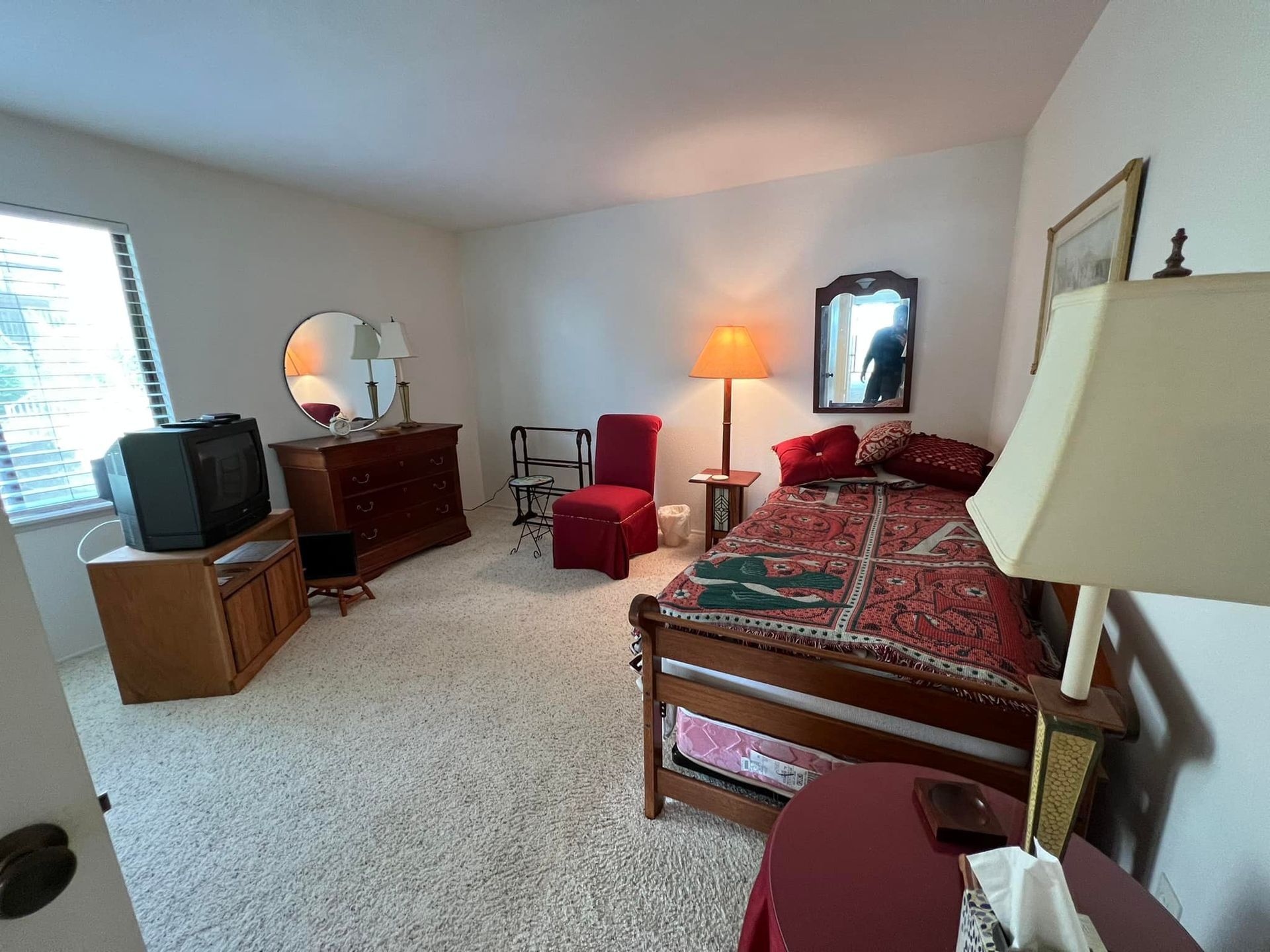 Bedroom with a twin bed, antique dresser with a mirror, a TV, and a red chair. The room has light-colored carpet and walls.