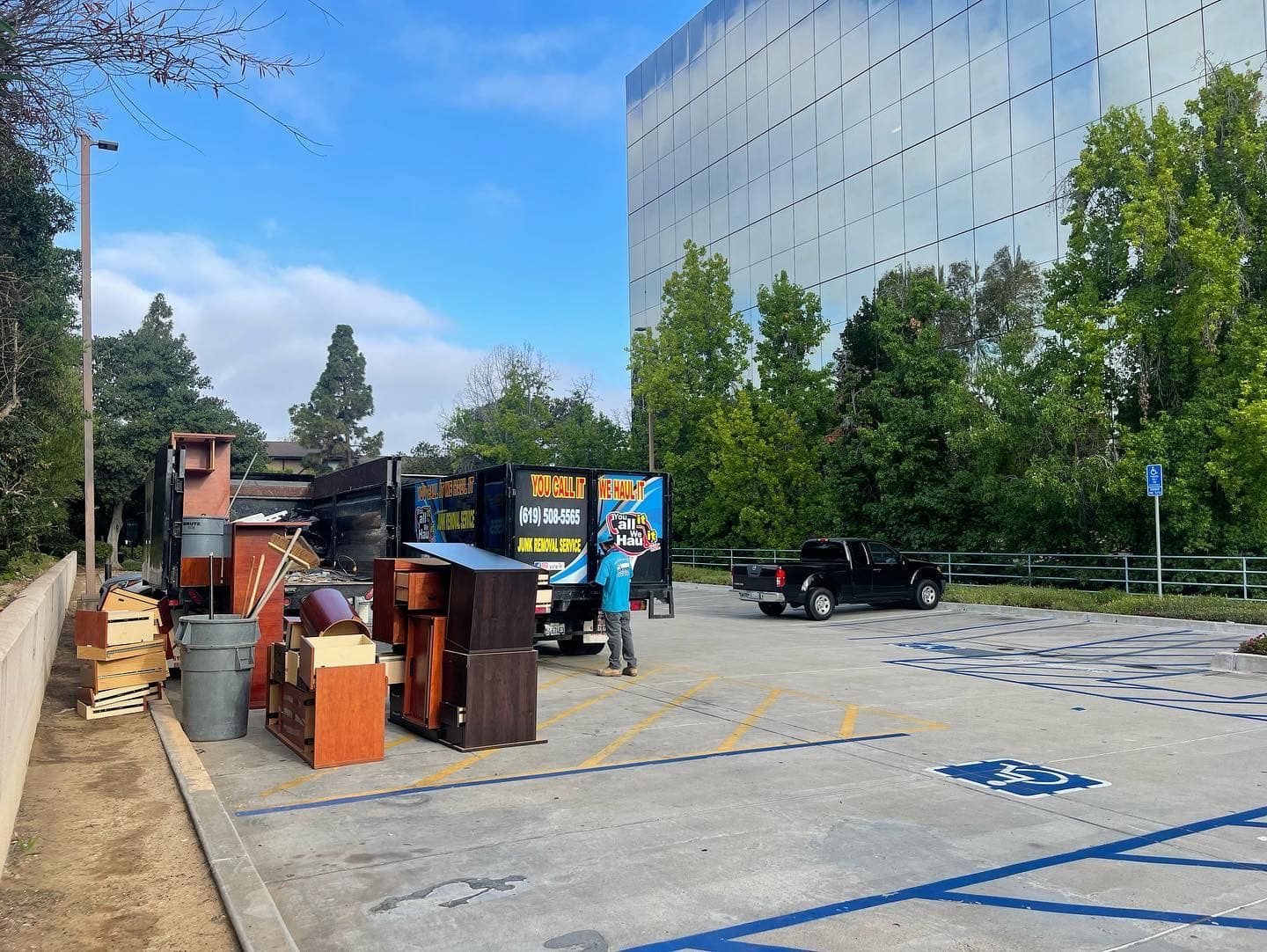 A parked black pickup truck and a junk removal truck with a worker. Items are loaded onto the truck in a parking lot.
