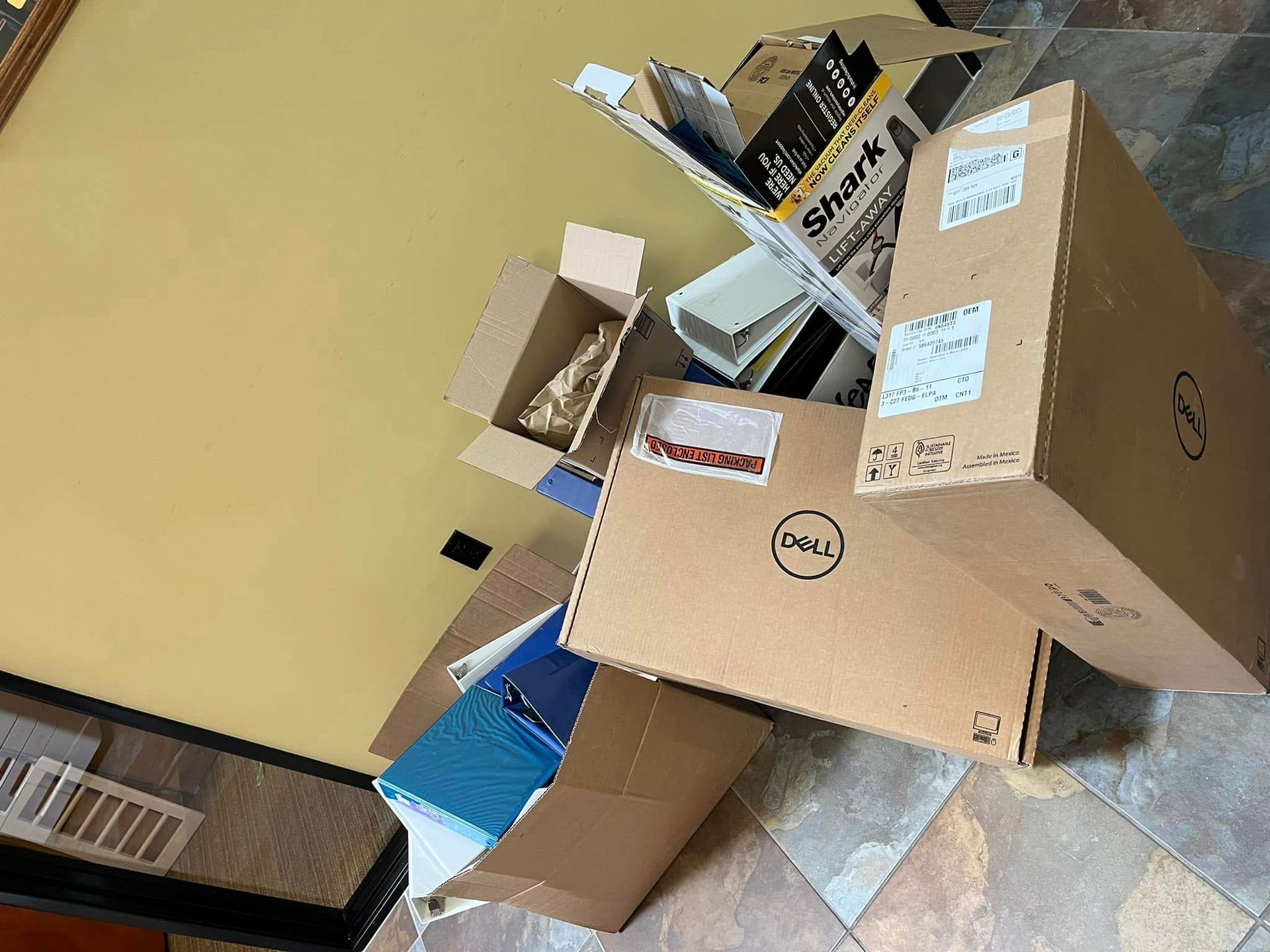 Pile of cardboard boxes, including a Dell box, on a tiled floor next to a light yellow wall.