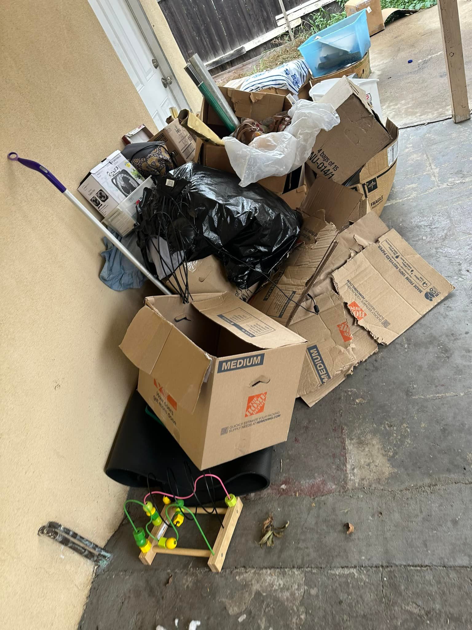 Pile of cardboard boxes, trash bags, and debris on a concrete floor, near a wall and doorway.