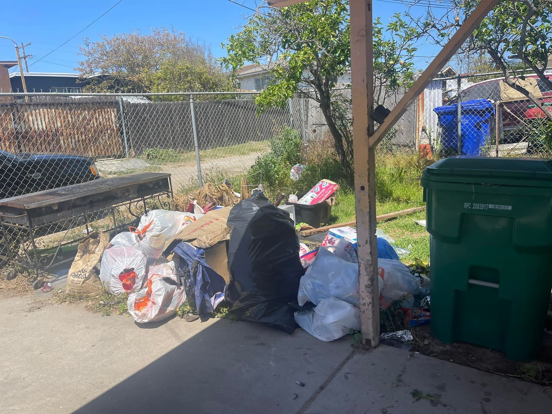 A pile of trash bags and a green trash bin sit next to a concrete area, next to a chain-link fence and overgrown foliage.