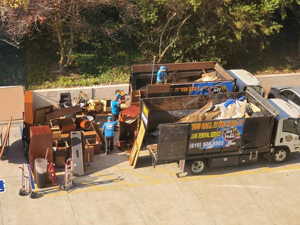 Workers loading debris into a dump truck. Piles of furniture and other items are on the ground. A sunny outdoor setting.