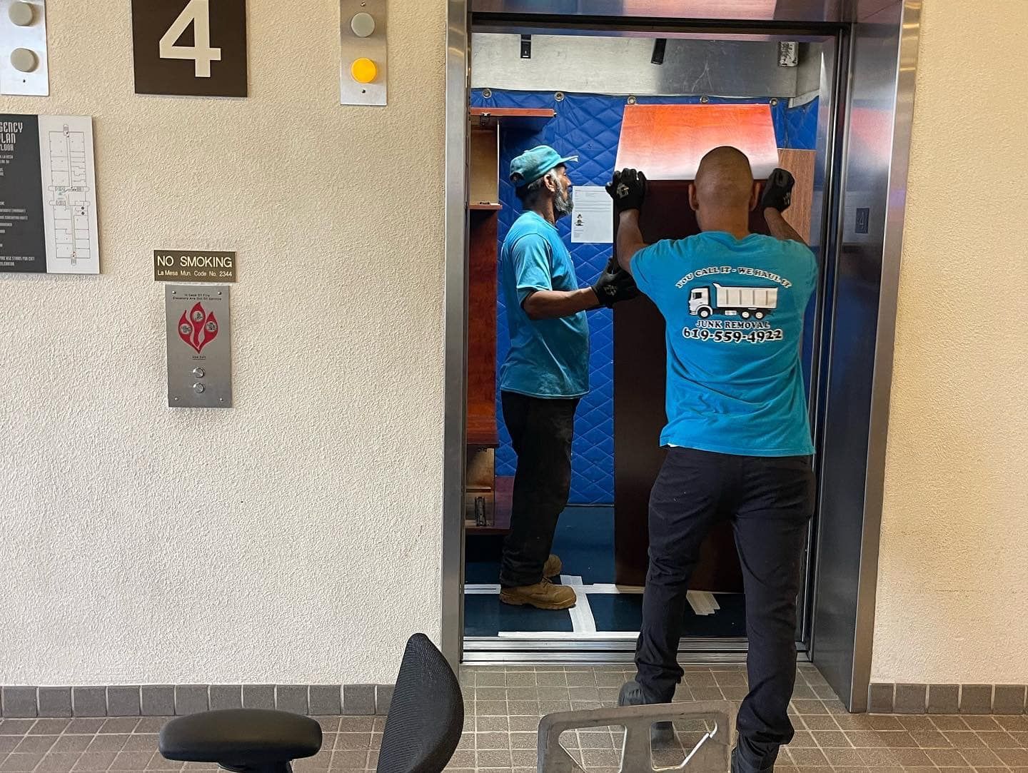 Two movers in blue shirts load an item into an elevator on the fourth floor. The elevator is open, and the hallway has a beige wall.