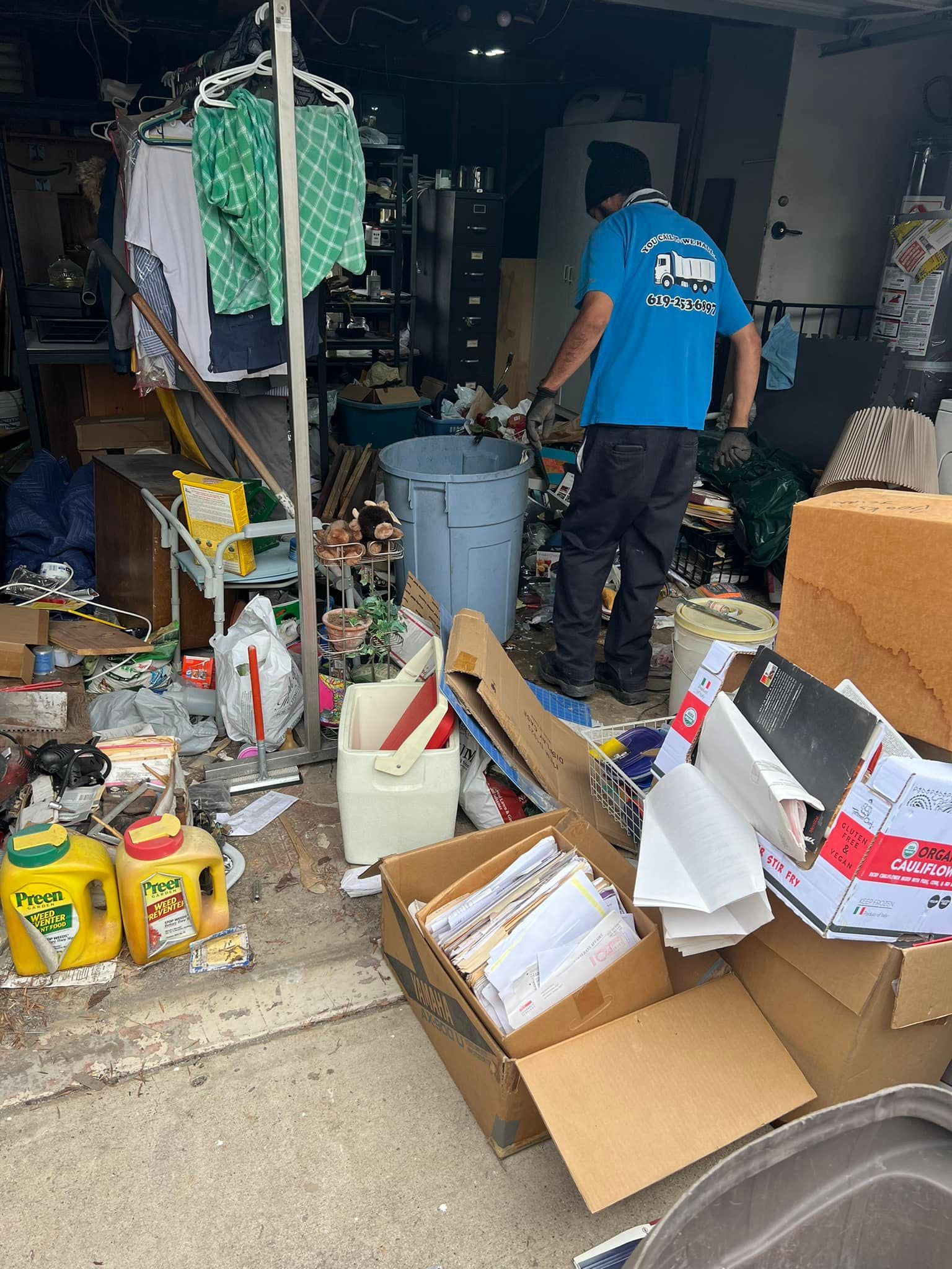 A person wearing a blue shirt stands amidst a cluttered garage, sorting through debris and trash. Various items and boxes are scattered about.