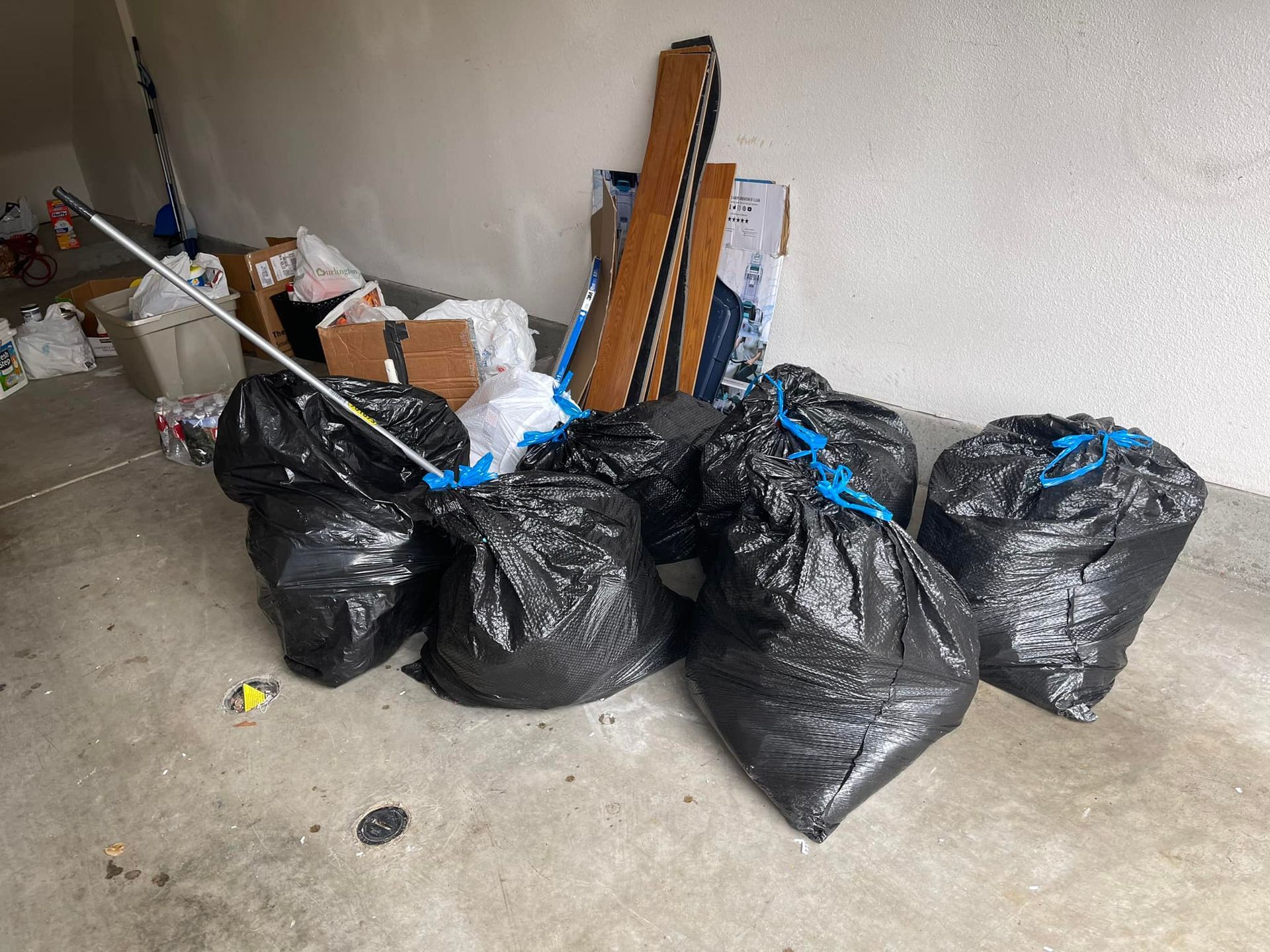 Five black trash bags tied with blue string sit on a concrete floor next to a pile of boxes and other debris.