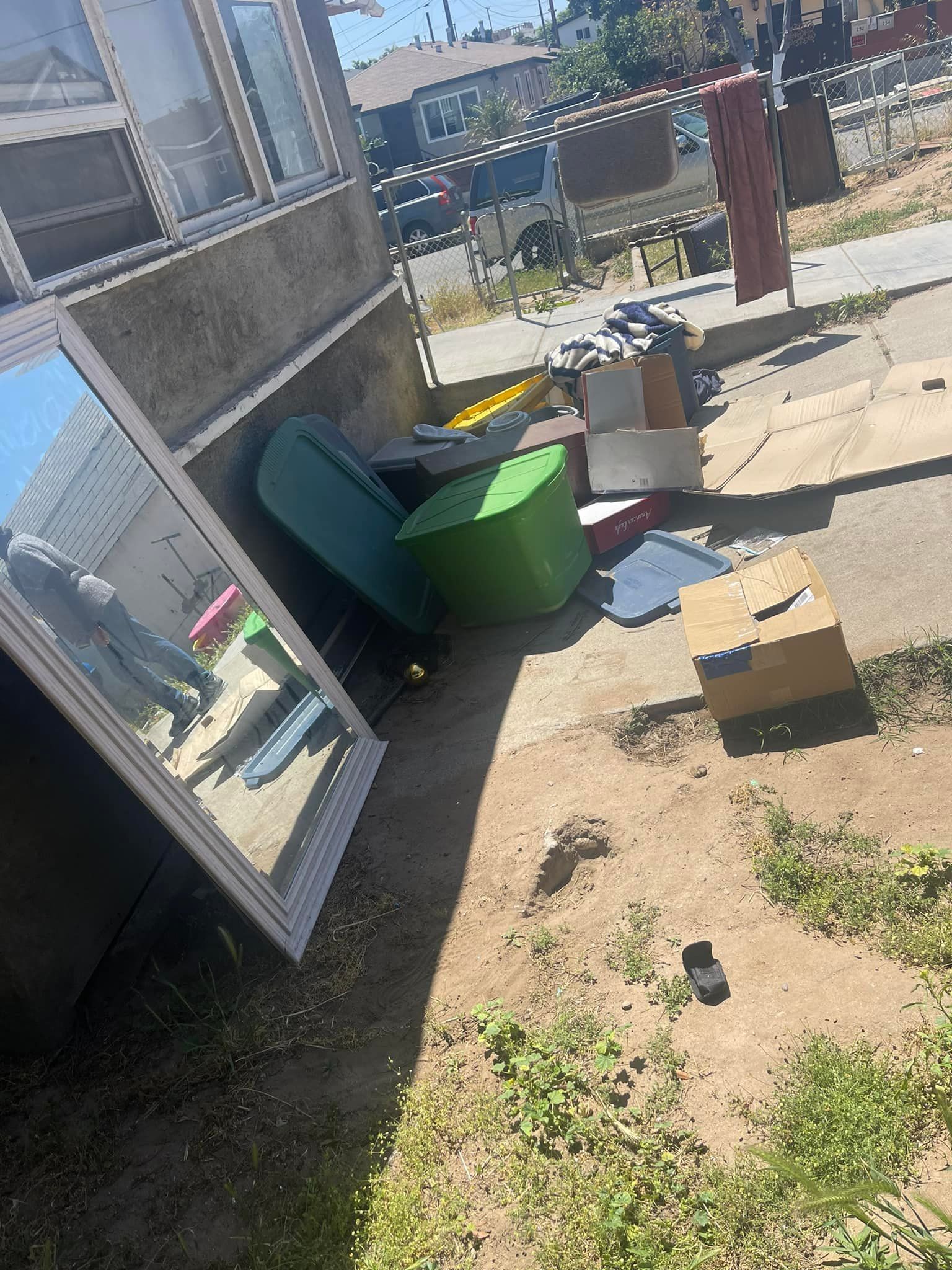Outside a building, discarded items including a mirror, bins, boxes, and various objects are piled on dirt and a sidewalk in the sunlight.