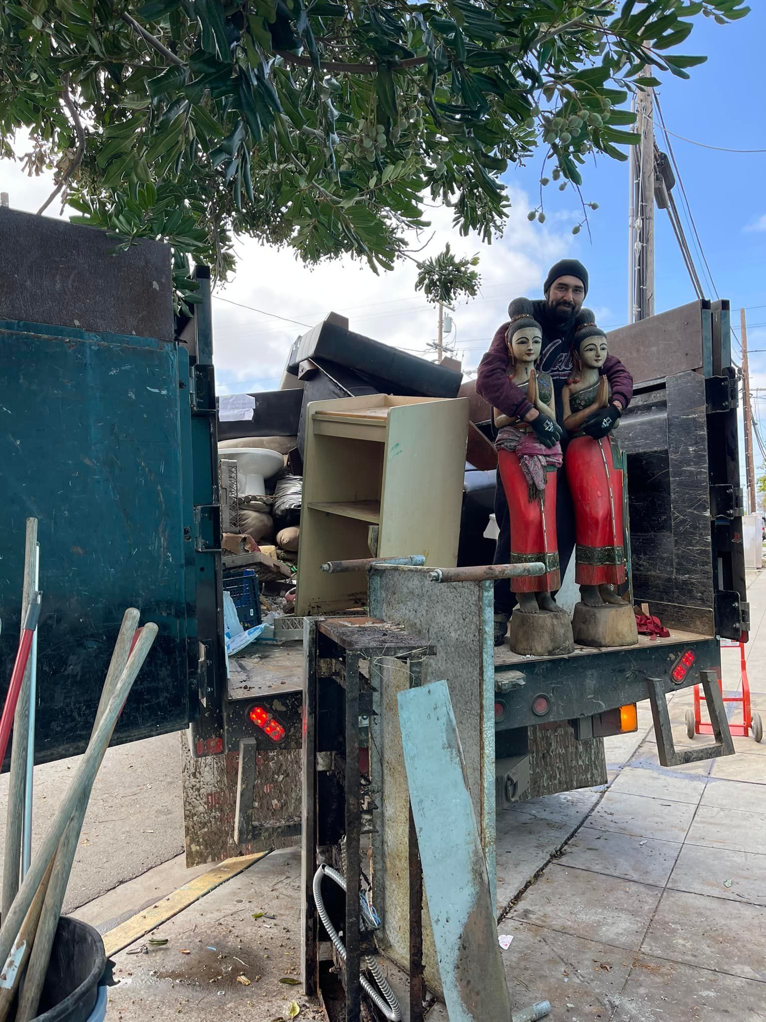 A person stands next to two statues in the back of a truck filled with debris. The statues are of people in red clothing.