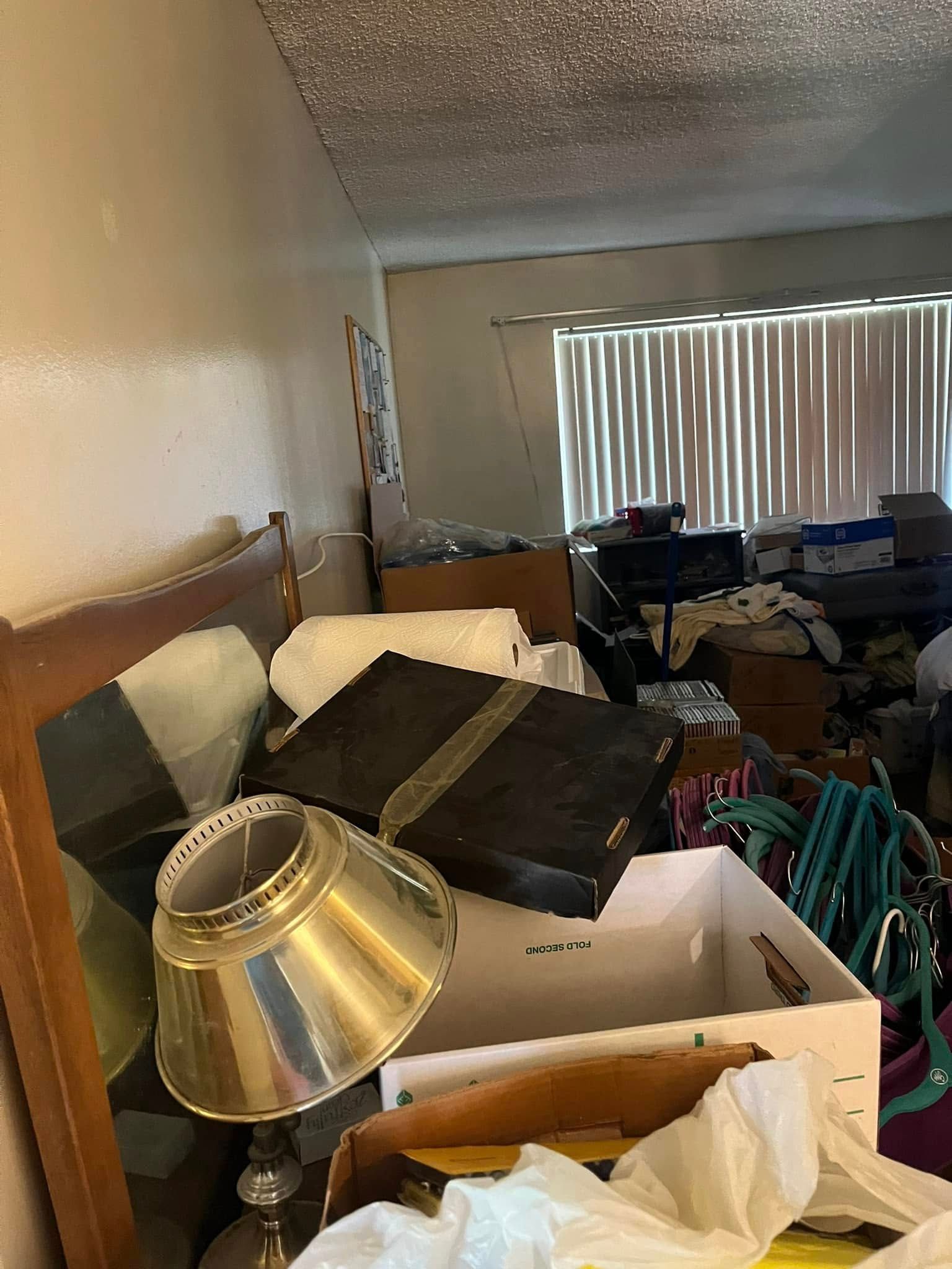 A cluttered room with various boxes, bags, and belongings near a bed and window with closed blinds. A lamp is in the foreground.