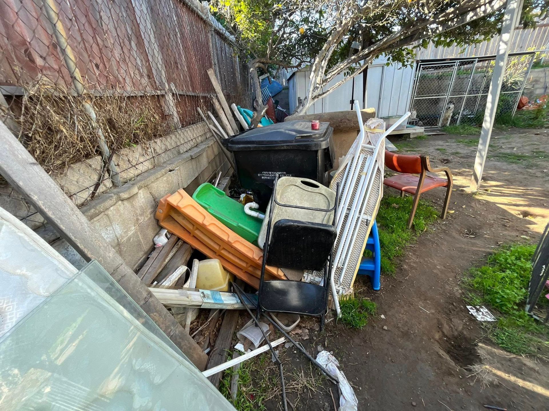 A cluttered outdoor area with a black trash can, broken furniture, and construction debris piled against a weathered fence.
