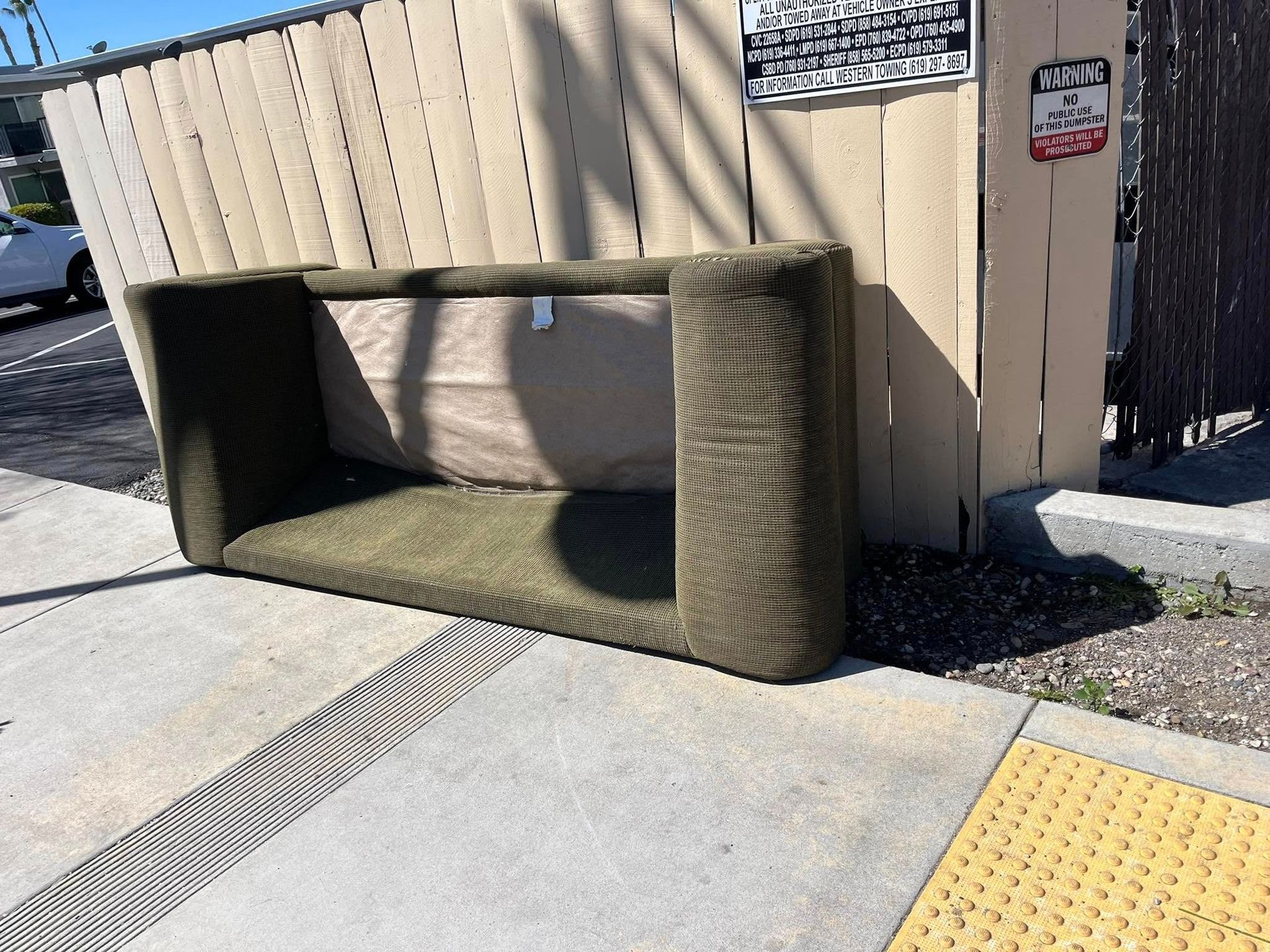 A worn green couch discarded on a sidewalk next to a wooden fence. The back of the couch is visible.