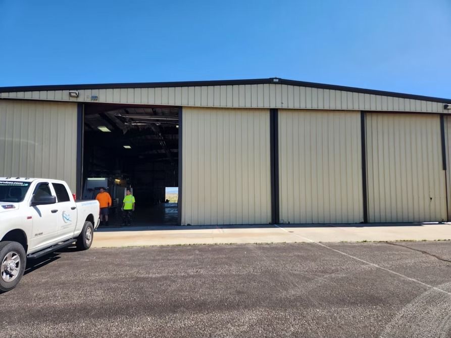 A white truck parked in front of a tan metal hangar with an open doorway. Several people in the hangar.