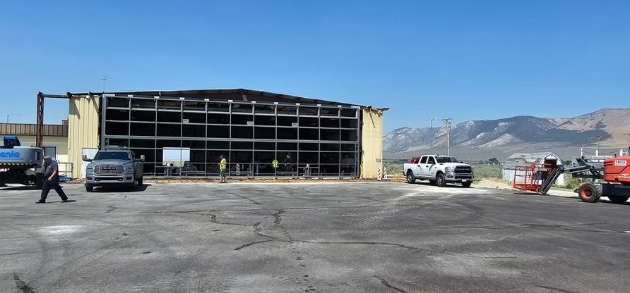 Building frame after a fire with vehicles parked in front on an asphalt surface, mountains in background.