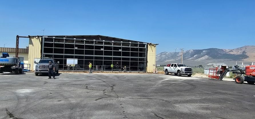 Building with exposed steel framework, trucks, workers in a lot, with mountains in the background under a blue sky.