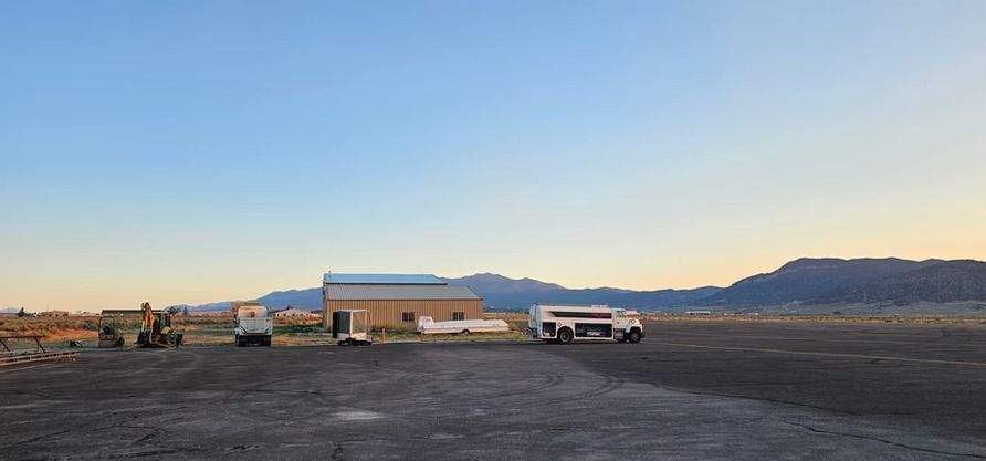 A rural landscape with a beige building, vehicles, and mountains under a blue sky.