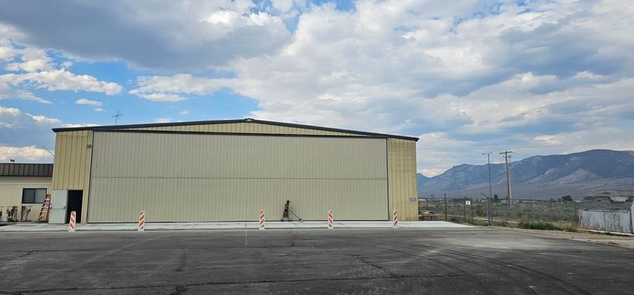 A large, tan hangar on a concrete pad. Orange traffic cones line the pad. Mountains are in the background under a cloudy sky.