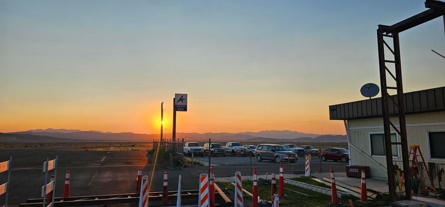 Sunset over an airfield with a building on the right, cars parked, and mountains in the distance.