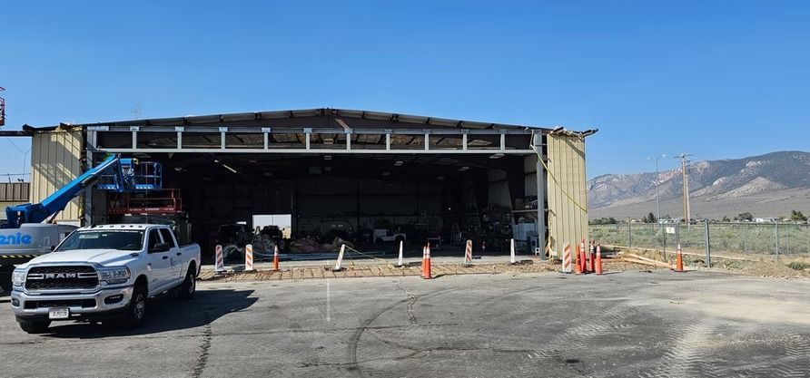 White truck parked in front of an open hangar with construction cones and a lift; mountains in the background.