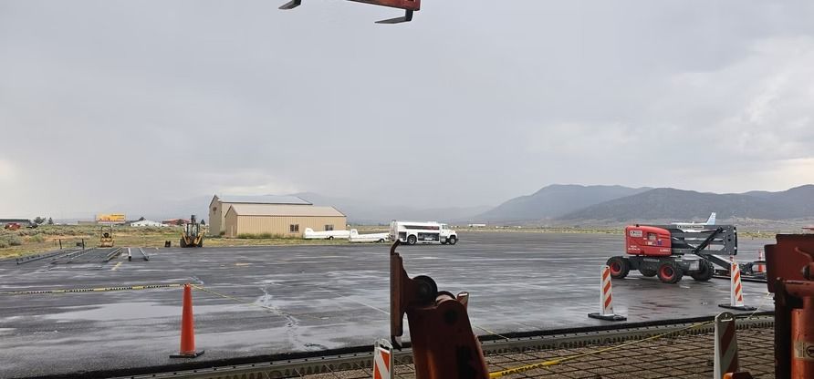 A wet airfield with a red tractor, a building, a white vehicle, mountains, and a cloudy sky.