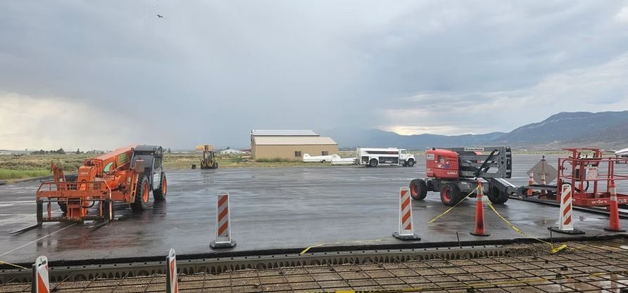 Construction site with heavy machinery, buildings, and mountains under cloudy sky. Wet pavement.