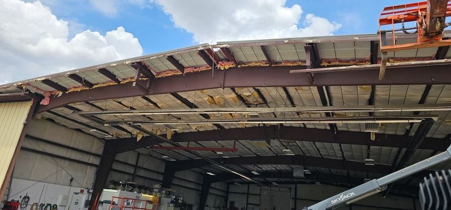 Damaged building roof with exposed steel beams and partial roof panels. Blue sky with clouds.