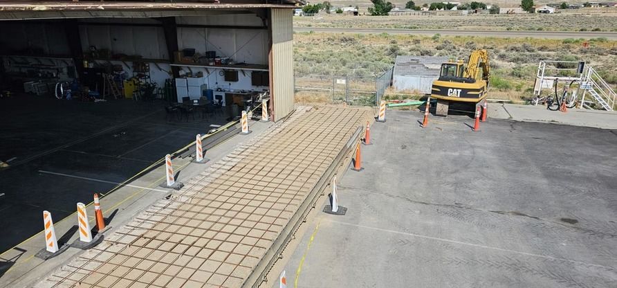 A paved ramp leads from a hangar. An excavator is parked near orange cones.