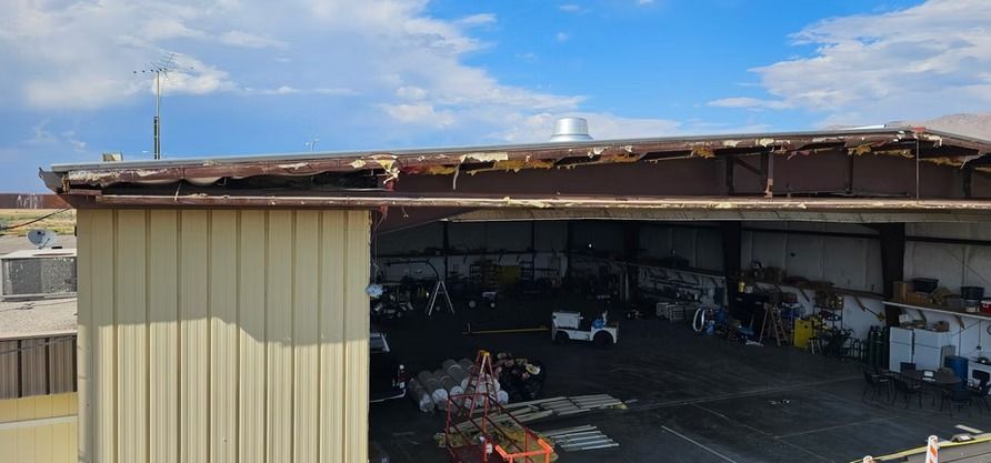 Exterior view of a metal building with a damaged roof and a partially visible interior containing equipment and objects.