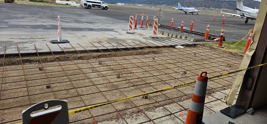 Airport construction site with rebar grid, orange cones, and airplanes in the background.