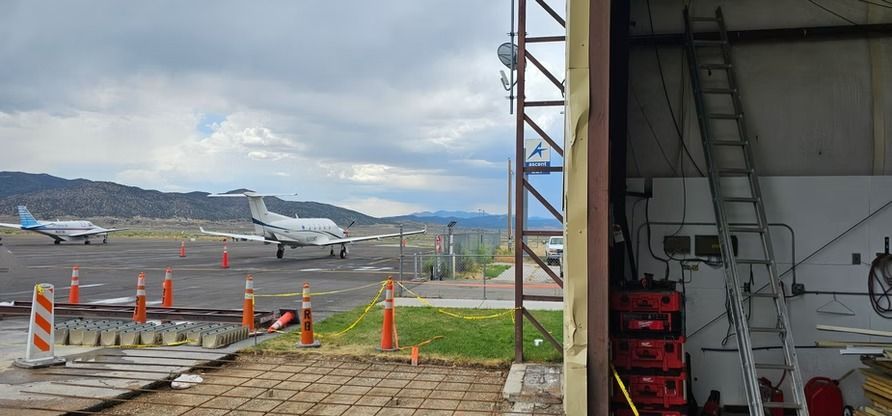 Airplanes on a runway, viewed from a hangar. Overcast sky, mountains in the distance. Construction in foreground.