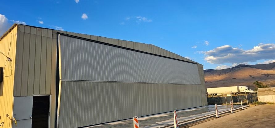 Large metal industrial building with a curved roof. Mountains and a blue sky in the background.