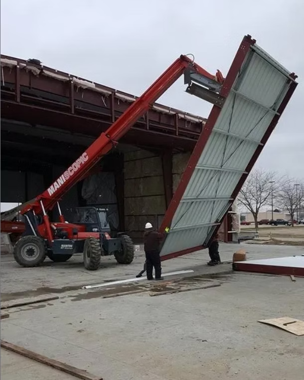 A telehandler lifts a large metal panel for construction. Two workers guide the panel. Gray overcast sky.