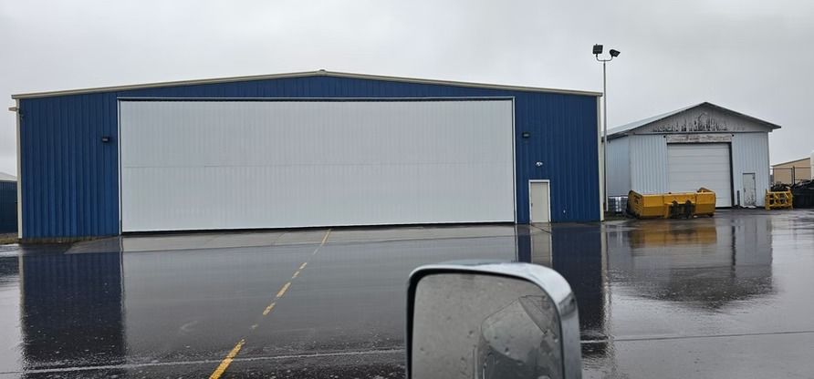 A large blue hangar with a white door, reflected in a wet asphalt lot on a cloudy day.