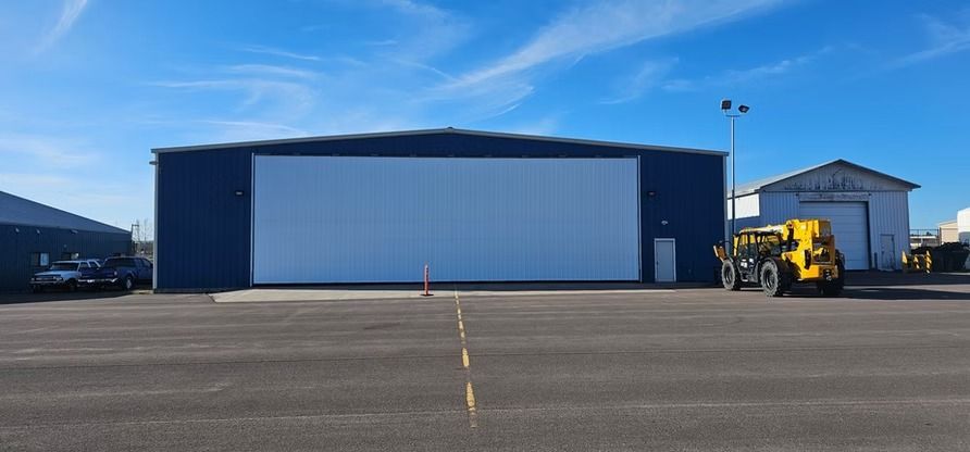 Large blue hangar with white door, on asphalt. A yellow tractor sits nearby, and a blue sky overhead.