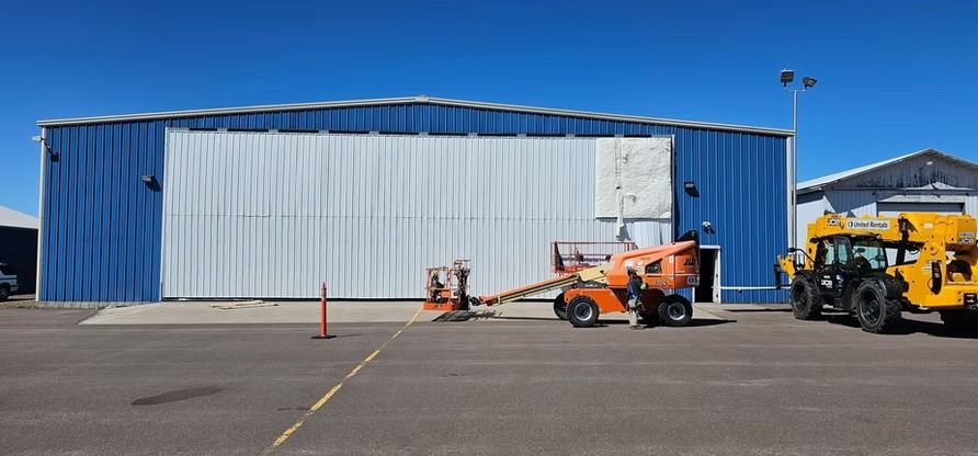 Blue and white hangar with orange lift equipment in front, a yellow loader to the right, and a clear blue sky.