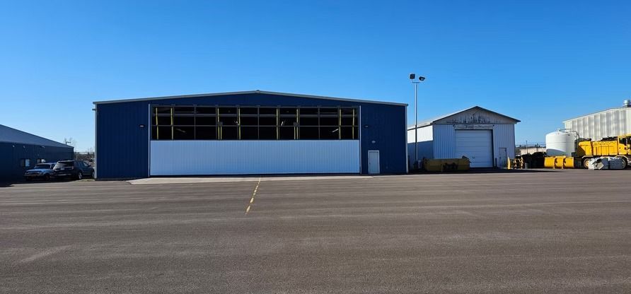 Blue hangar with white lower section and a small adjacent building on a paved surface under a clear blue sky.
