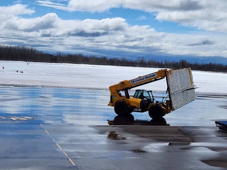 Yellow forklift carrying a large reflective panel on a wet surface with snow and trees in the background.