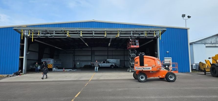 Blue hangar with open door, orange lift in foreground, people working.