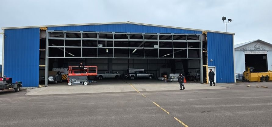 Large blue hangar with open doors, vehicles and people visible inside and outside, gray sky.