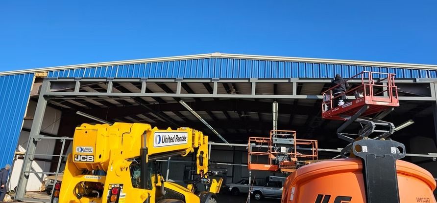 Construction site with a blue roof, metal beams, and heavy machinery, including a yellow forklift and orange lifts.