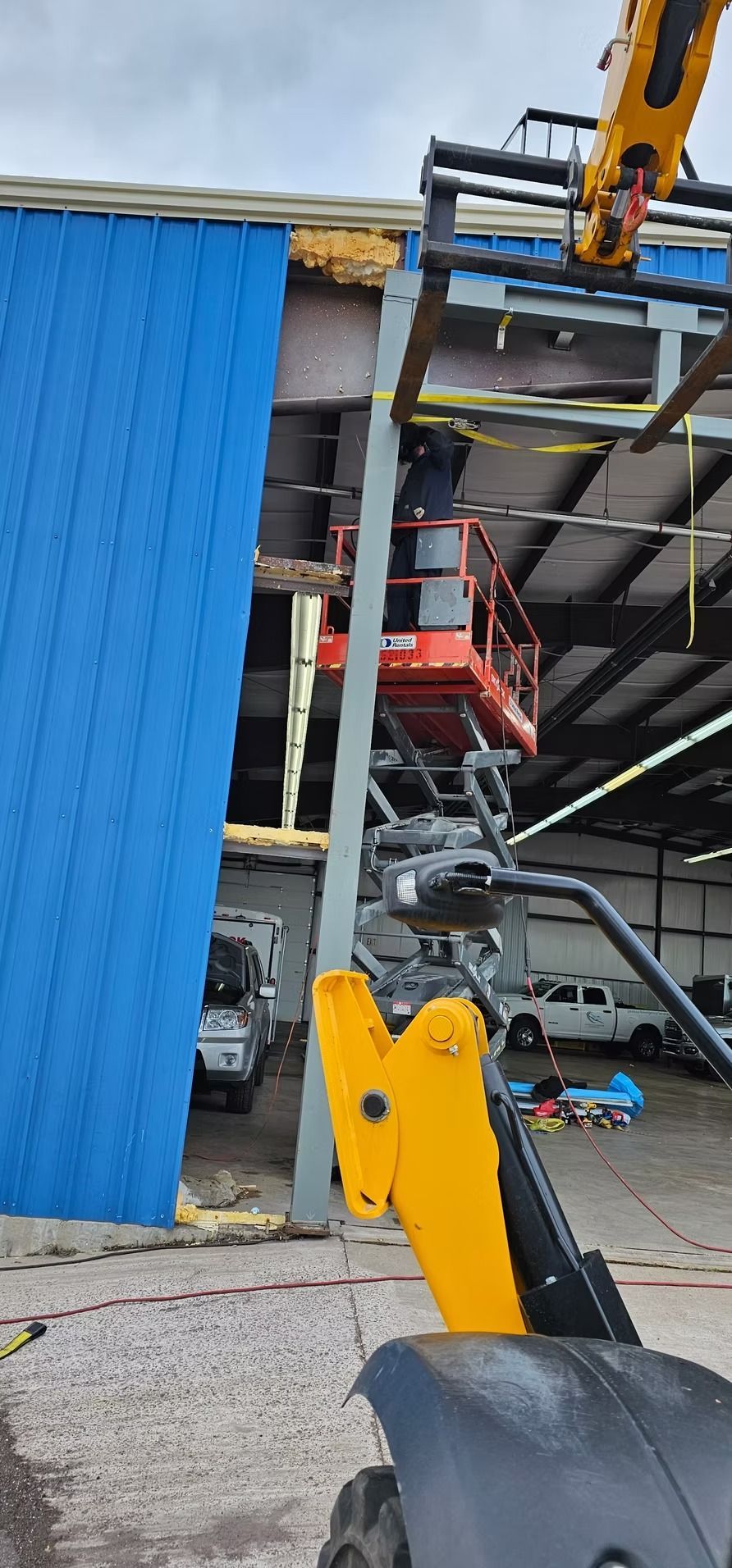 Construction worker on a lift near a blue building with yellow machinery in the foreground.