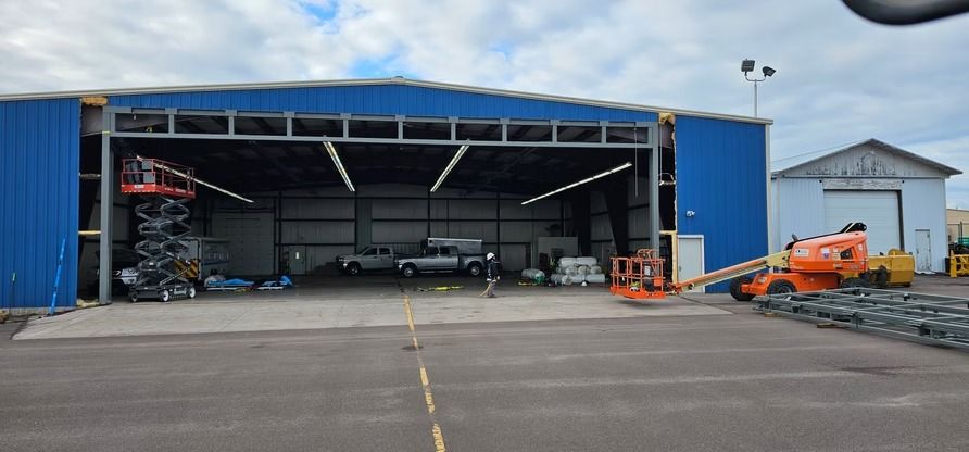 Blue hangar with open door, vehicles inside and a lift. Another building and lift outside. Paved area.