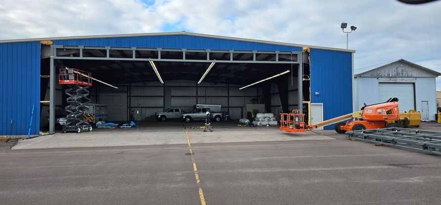A blue hangar with an open door, trucks inside, and construction equipment outside.