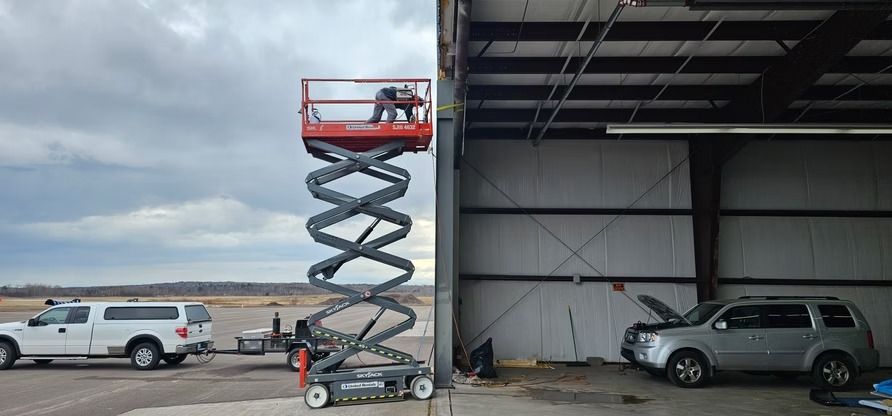 Person on a scissor lift near a building's open doorway. A truck and SUV are nearby. Overcast sky.