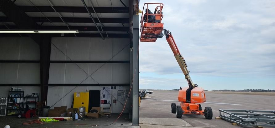 Person in an orange lift working on a metal building. The lift is on a paved surface. Cloudy sky.