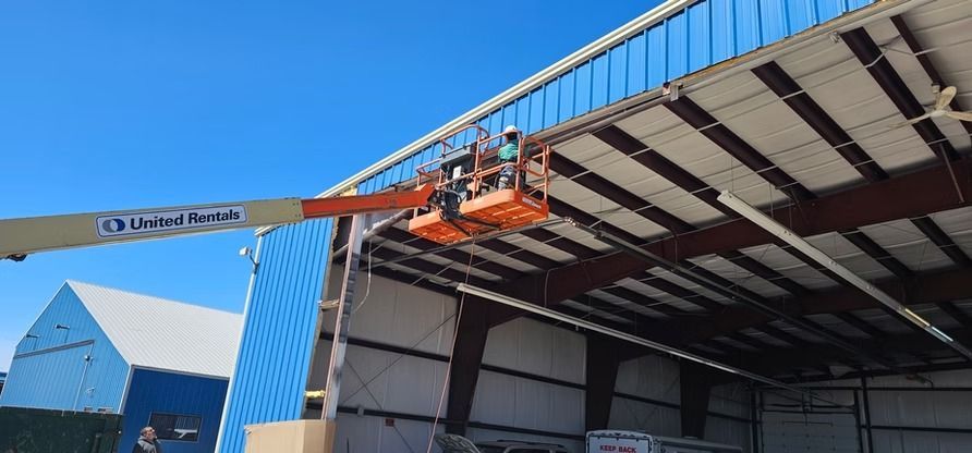 Workers in an orange lift platform near blue roof of a large building. Bright sky.