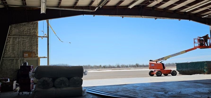 Inside a warehouse, a person operates an orange lift; the open door reveals a blue sky and stacks of green boards.