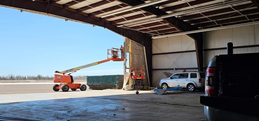 Orange lift extending from a hangar opening. A person is in the lift's bucket. A white SUV is parked inside.