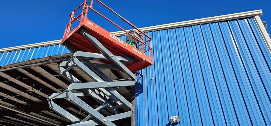 A scissor lift extended next to a blue metal building under a bright blue sky.