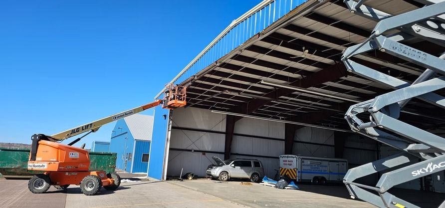 A boom lift is extended toward a blue building to access the roof. A scissor lift is on the right side.